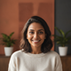 Close-up portrait of a smiling South Asian woman in her late 20s, looking at the camera. Softly blurred background with warm 